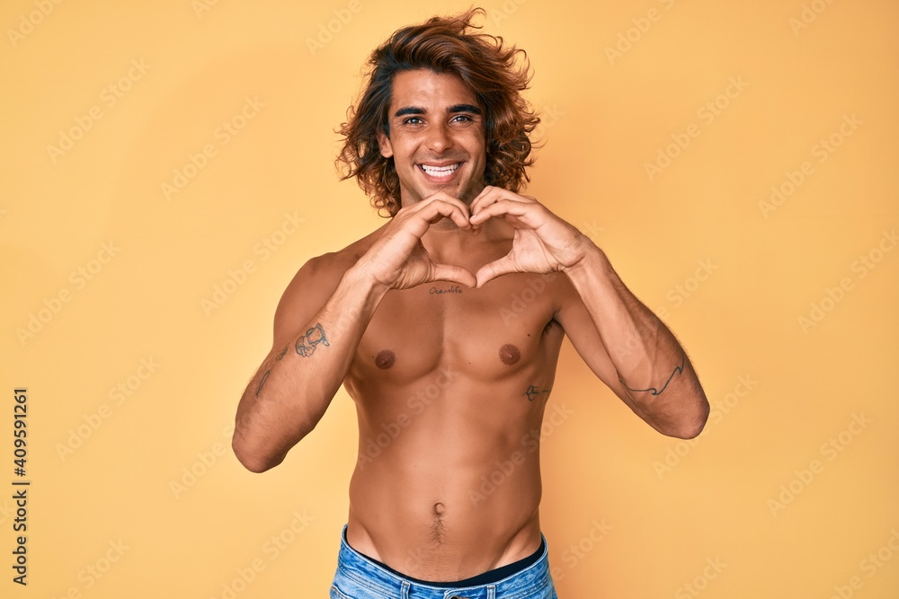 Young hispanic man standing shirtless smiling in love doing heart symbol shape with hands. romantic concept.