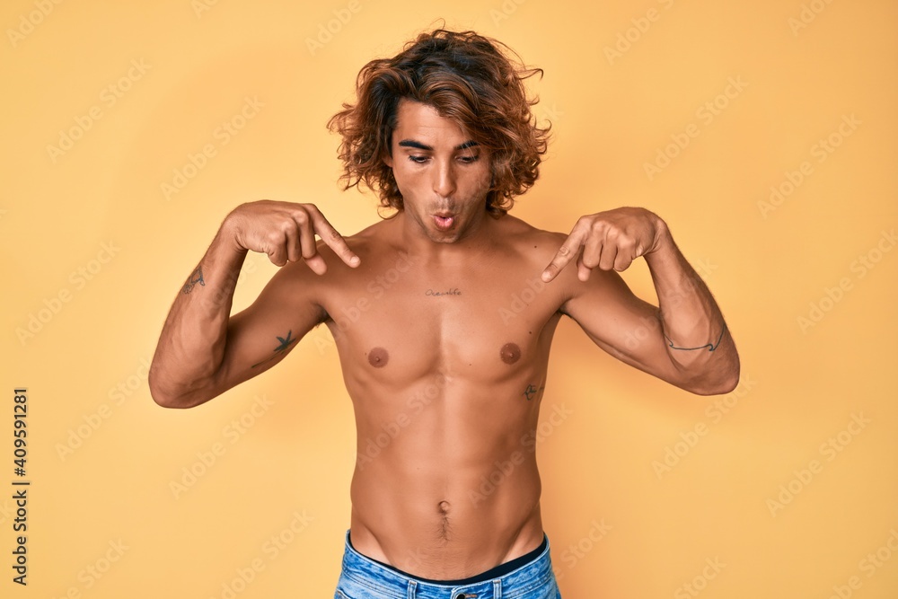 Young hispanic man standing shirtless pointing down with fingers showing advertisement, surprised face and open mouth