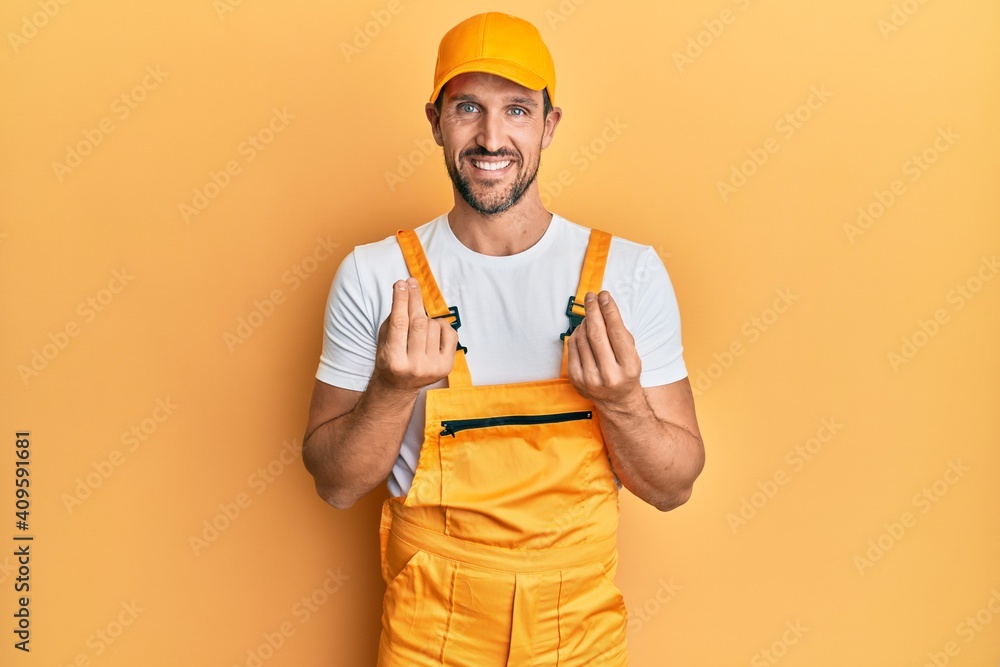 Young handsome man wearing handyman uniform over yellow background doing money gesture with hands, asking for salary payment, millionaire business - obrazy, fototapety, plakaty 
