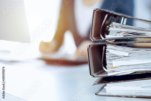 Binders with papers are waiting to be processed by business woman or bookkeeper working at the desk in office back in blur. Audit and tax concept