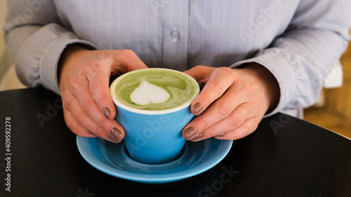 Woman holding blue cup with hot trendy green latte with art heart on the foam on black wooden table background  healthy drink with latte art
