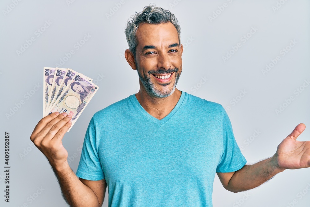 Middle age grey-haired man holding japanese yen banknotes celebrating achievement with happy smile and winner expression with raised hand Middle age grey-haired man holding japanese yen banknotes celebrating achievement with happy smile and winner expression with raised hand