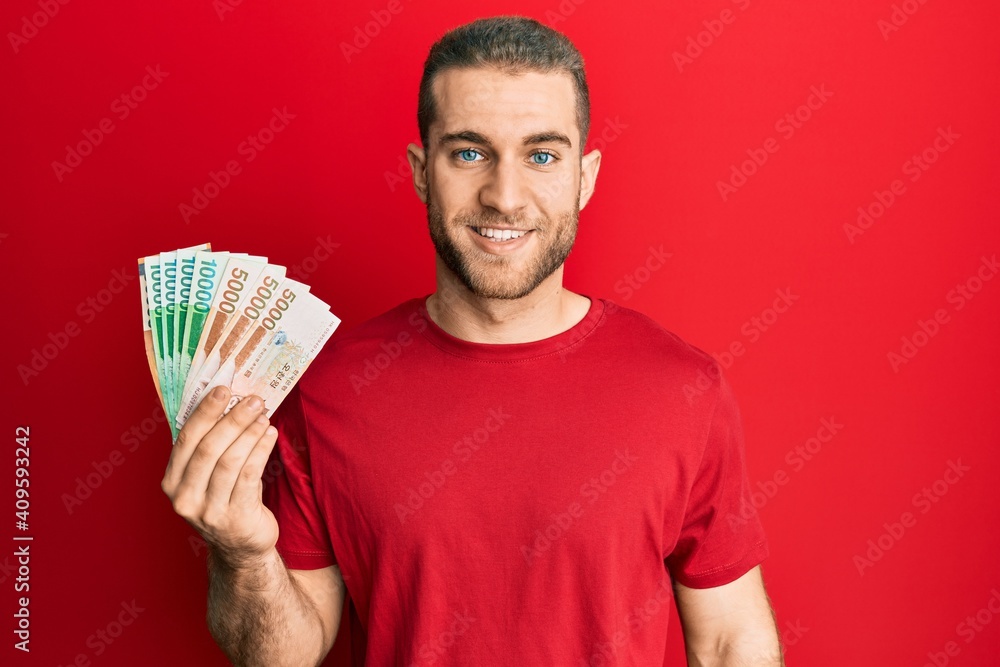 Young caucasian man holding 5000 south korean won banknotes looking positive and happy standing and smiling with a confident smile showing teeth