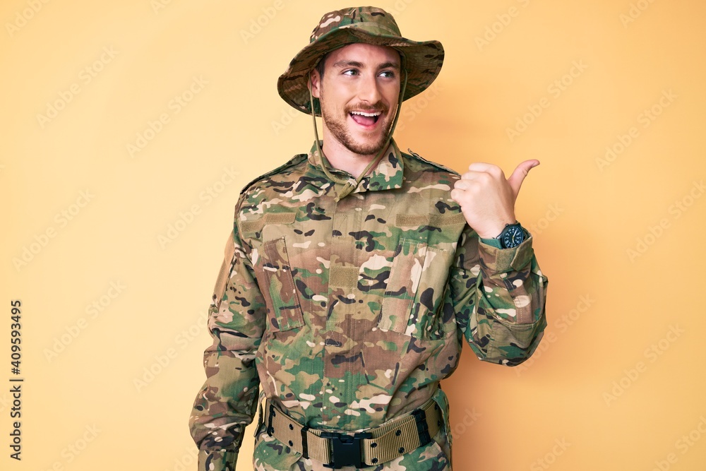 Young caucasian man wearing camouflage army uniform smiling with happy face looking and pointing to the side with thumb up. Young caucasian man wearing camouflage army uniform smiling with happy face looking and pointing to the side with thumb up.