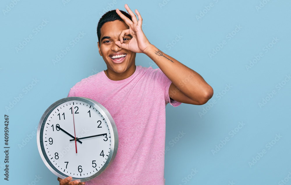Young handsome hispanic man holding big clock smiling happy doing ok sign with hand on eye looking through fingers
