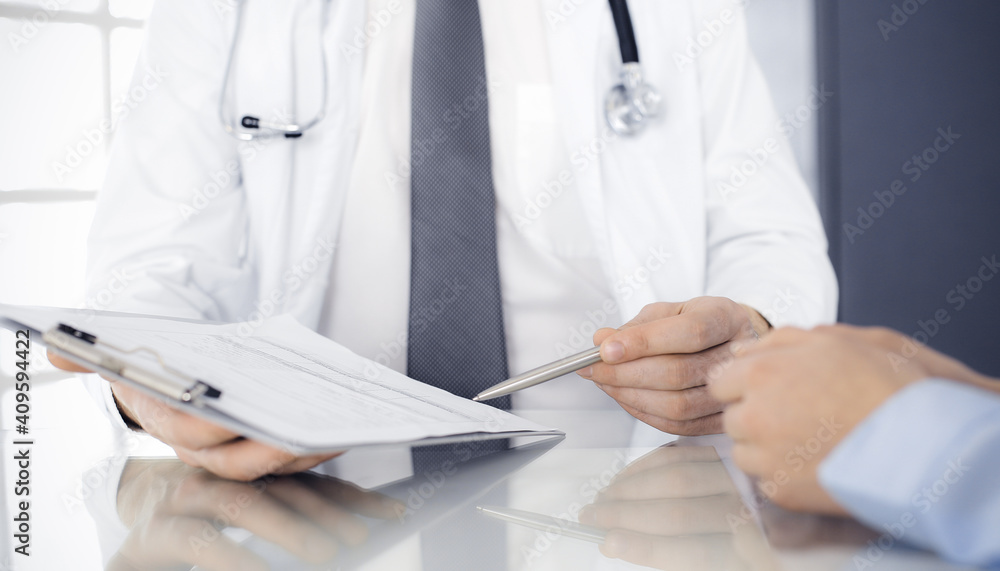 Unknown male doctor and woman-patient discussing current health examination while sitting in clinic and using clipboard. Good medical service in hospital. Medicine concept Unknown male doctor and woman-patient discussing current health examination while sitting in clinic and using clipboard. Good medical service in hospital. Medicine concept