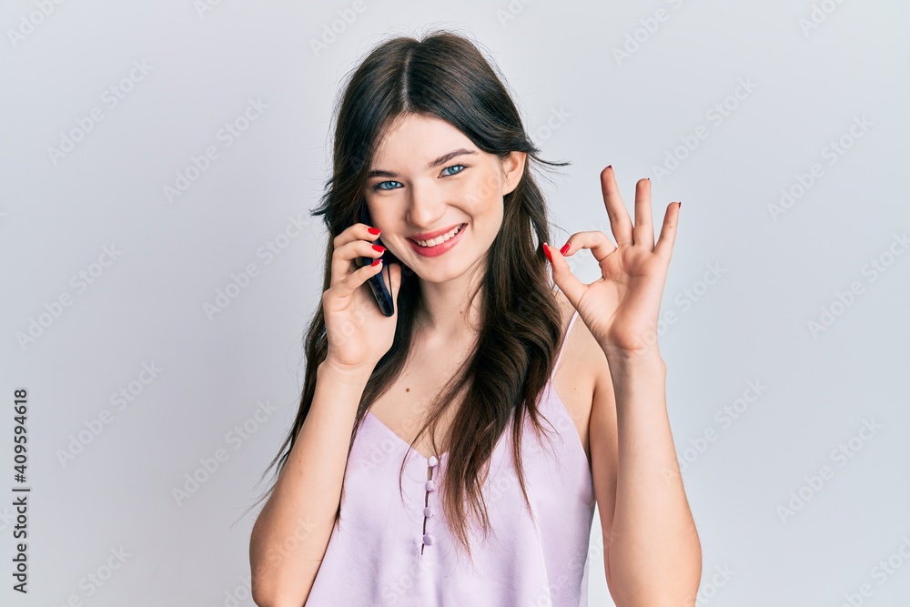 Young beautiful caucasian girl having conversation talking on the smartphone doing ok sign with fingers, smiling friendly gesturing excellent symbol Young beautiful caucasian girl having conversation talking on the smartphone doing ok sign with fingers, smiling friendly gesturing excellent symbol