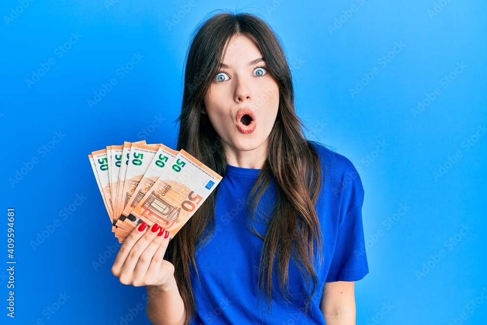 Young beautiful caucasian girl holding bunch of 50 euro banknotes scared and amazed with open mouth for surprise, disbelief face Young beautiful caucasian girl holding bunch of 50 euro banknotes scared and amazed with open mouth for surprise, disbelief face
