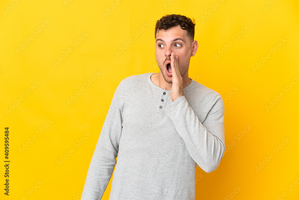 Young caucasian handsome man isolated on yellow background whispering something with surprise gesture while looking to the side - obrazy, fototapety, plakaty 