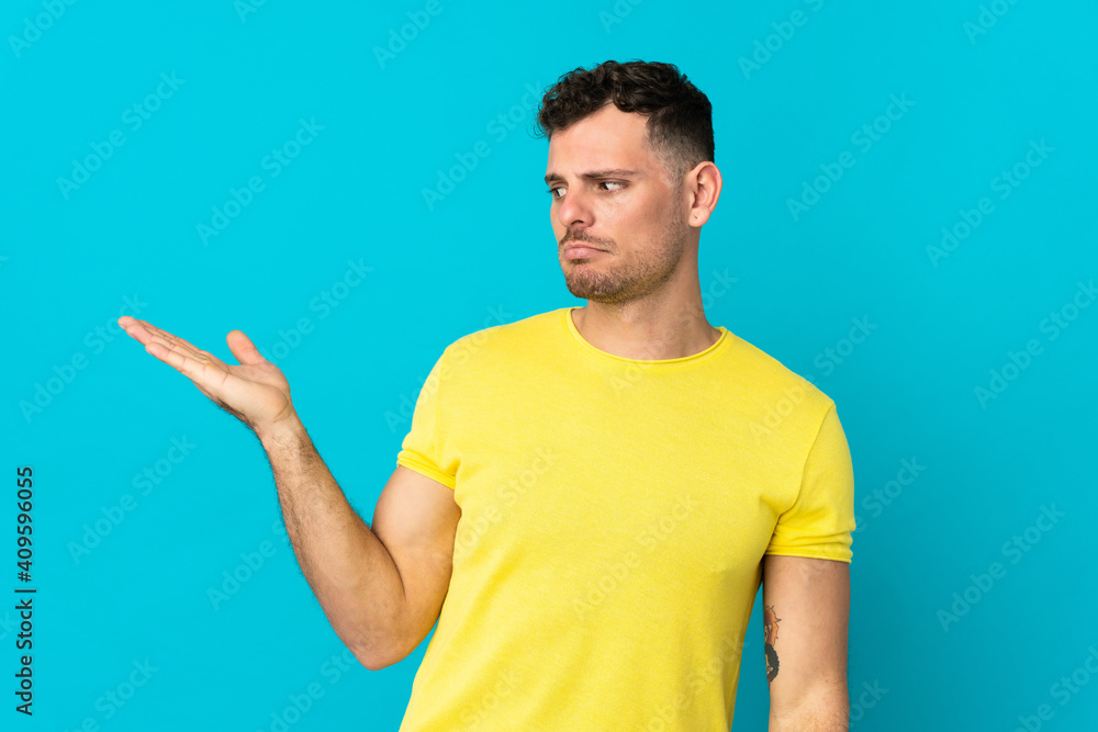 Young caucasian handsome man isolated on blue background holding copyspace with doubts