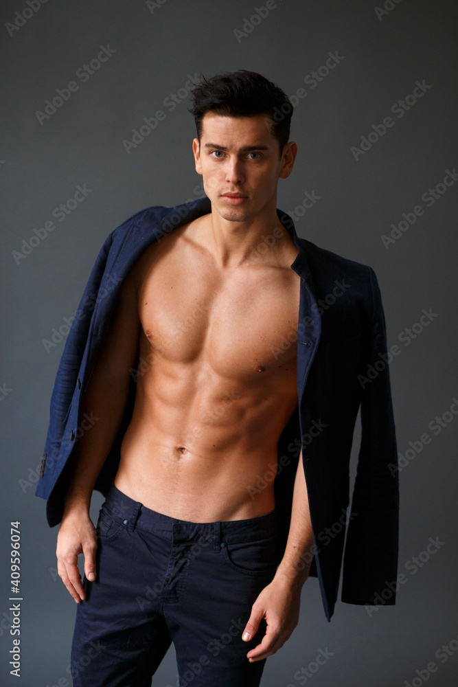 Elegant, muscular, handsome brunette man sitting and posing in fashionable suit on a naked torso, over grey background.