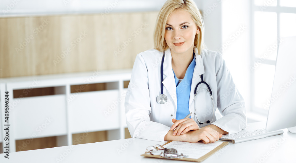 Portrate of woman-doctor at work while sitting at the desk in clinic. Blonde cheerful physician ready to help patients Portrate of woman-doctor at work while sitting at the desk in clinic. Blonde cheerful physician ready to help patients