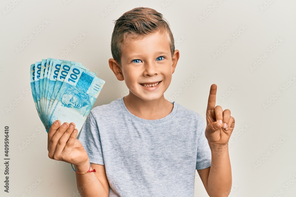 Adorable caucasian kid holding 100 brazilian real banknotes smiling with an idea or question pointing finger with happy face, number one Adorable caucasian kid holding 100 brazilian real banknotes smiling with an idea or question pointing finger with happy face, number one
