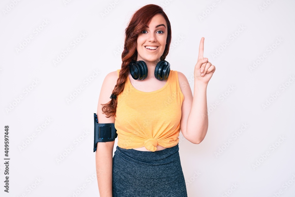 Young beautiful woman wearing gym clothes and using headphones pointing finger up with successful idea. exited and happy. number one. Young beautiful woman wearing gym clothes and using headphones pointing finger up with successful idea. exited and happy. number one.