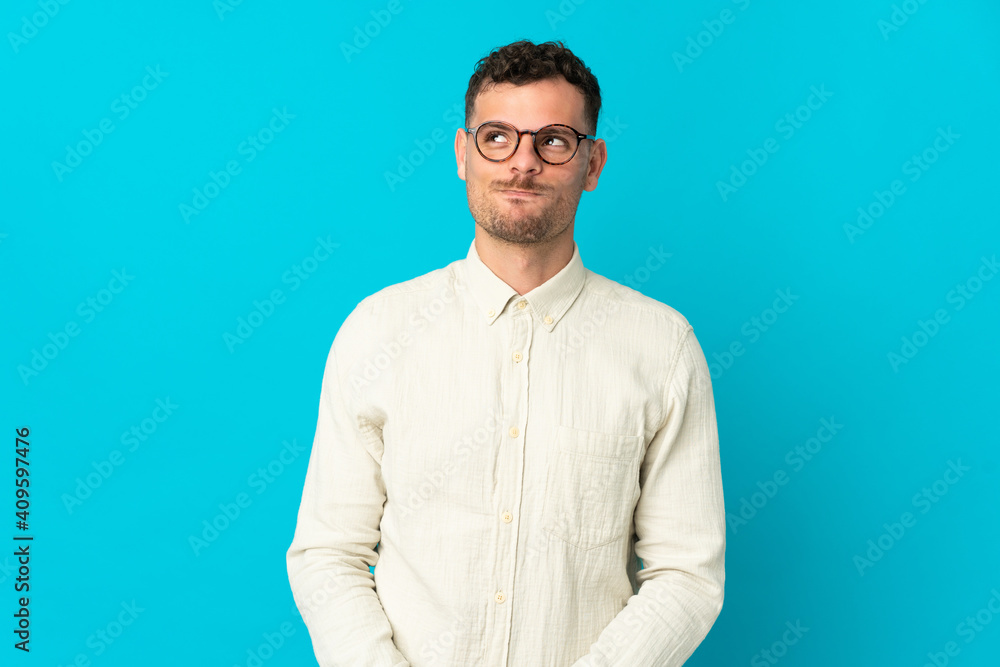 Young caucasian handsome man isolated on blue background having doubts while looking up Young caucasian handsome man isolated on blue background having doubts while looking up