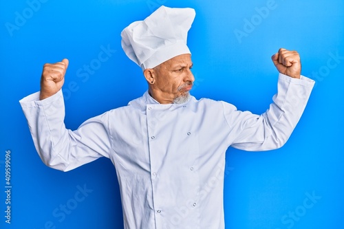 Middle age grey-haired man wearing professional cook uniform and hat showing arms muscles smiling proud. fitness concept.