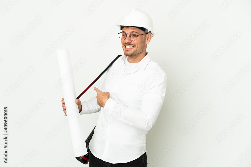 Young architect man with helmet and holding blueprints isolated on white background pointing back