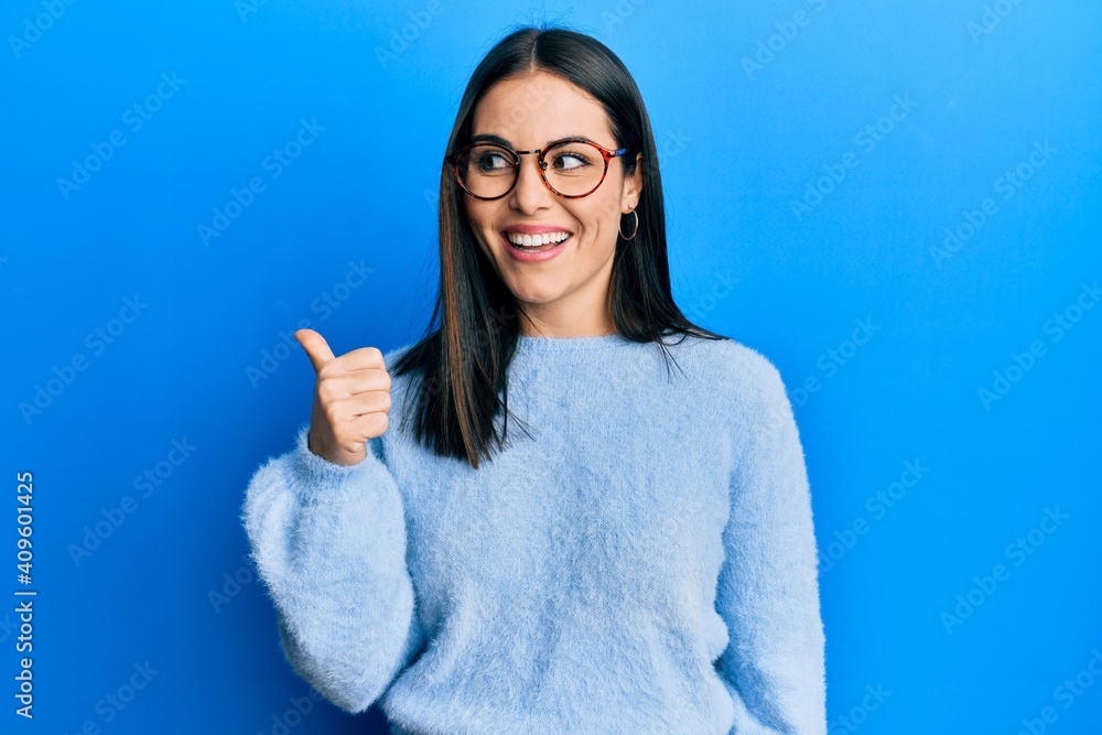 Young brunette woman wearing casual clothes and glasses pointing thumb up to the side smiling happy with open mouth Young brunette woman wearing casual clothes and glasses pointing thumb up to the side smiling happy with open mouth