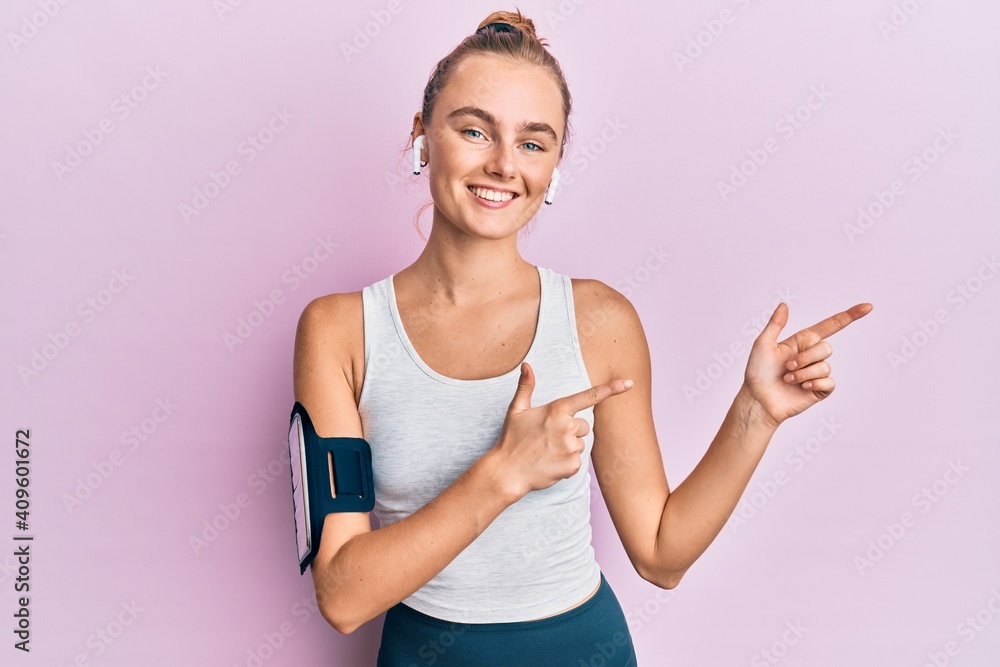 Beautiful blonde sport woman wearing arm band and earphones smiling and looking at the camera pointing with two hands and fingers to the side.