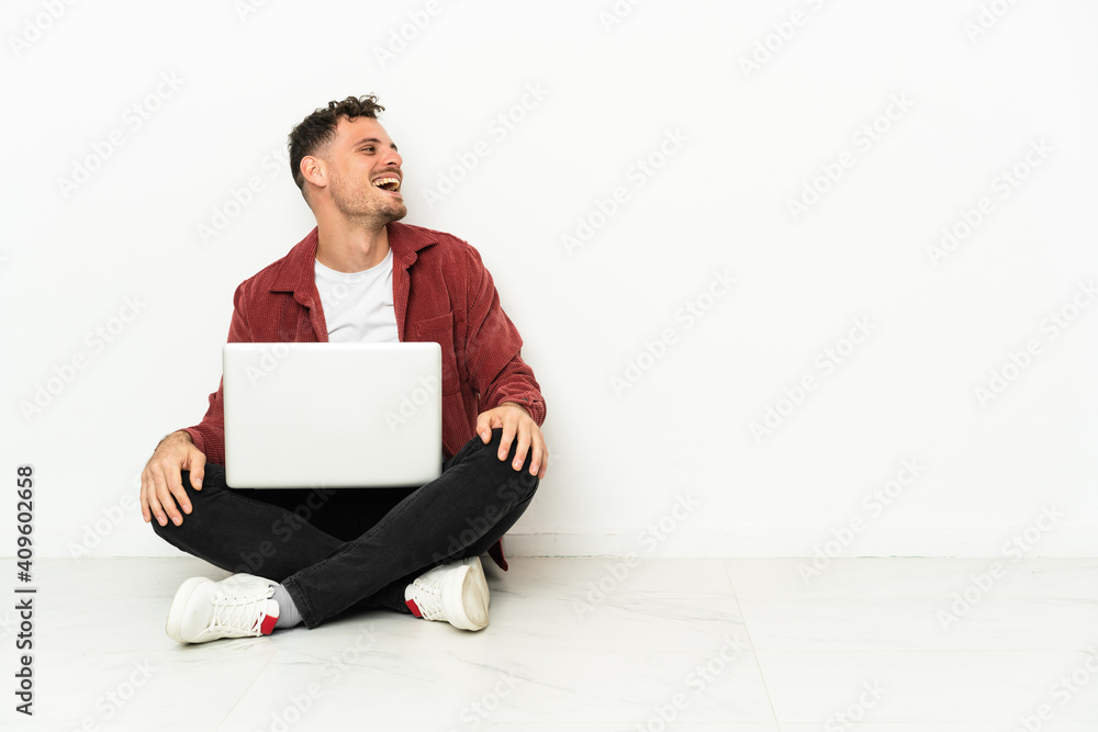 Young handsome caucasian man sit-in on the floor with laptop laughing in lateral position Young handsome caucasian man sit-in on the floor with laptop laughing in lateral position