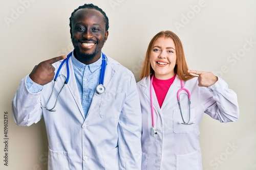 Young interracial couple wearing doctor uniform and stethoscope smiling cheerful showing and pointing with fingers teeth and mouth. dental health concept.