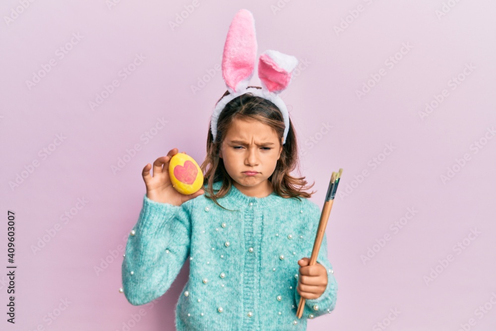 Little beautiful girl wearing cute easter bunny ears holding colored egg skeptic and nervous, frowning upset because of problem. negative person. Little beautiful girl wearing cute easter bunny ears holding colored egg skeptic and nervous, frowning upset because of problem. negative person.