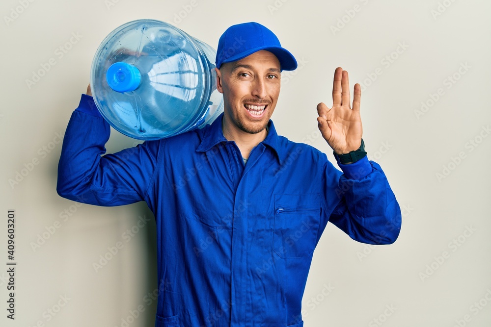 Bald courier man with beard holding a gallon bottle of water for delivery doing ok sign with fingers, smiling friendly gesturing excellent symbol