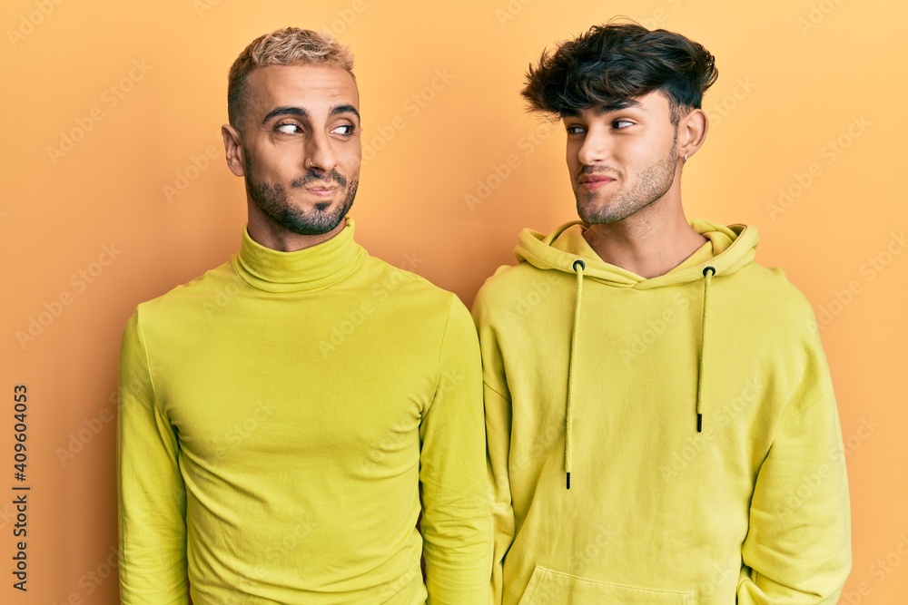 Obraz premium Homosexual gay couple standing together wearing yellow clothes smiling looking to the side and staring away thinking.
