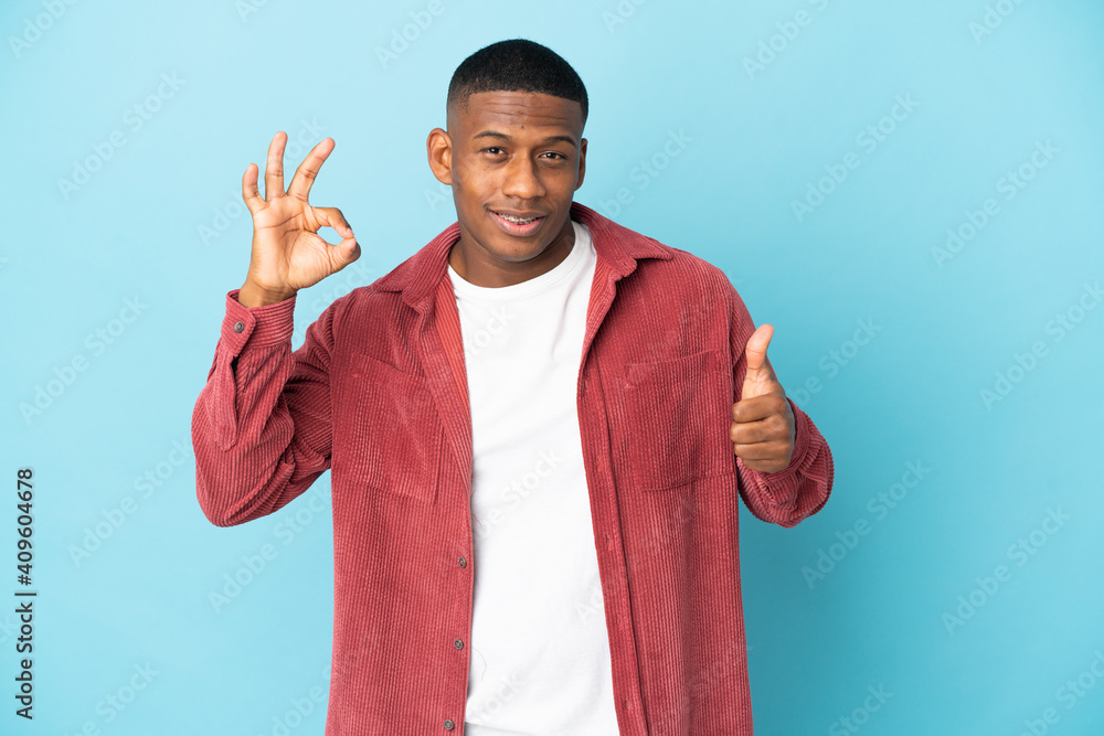 Young latin man isolated on blue background showing ok sign and thumb up gesture Young latin man isolated on blue background showing ok sign and thumb up gesture