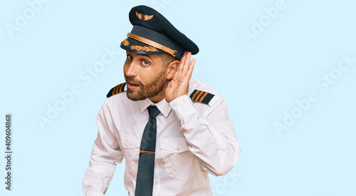 Handsome man with beard wearing airplane pilot uniform smiling with hand over ear listening an hearing to rumor or gossip. deafness concept. © Krakenimages.com
