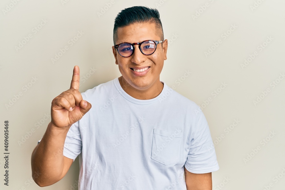 Young latin man wearing casual clothes and glasses smiling with an idea or question pointing finger up with happy face, number one Young latin man wearing casual clothes and glasses smiling with an idea or question pointing finger up with happy face, number one