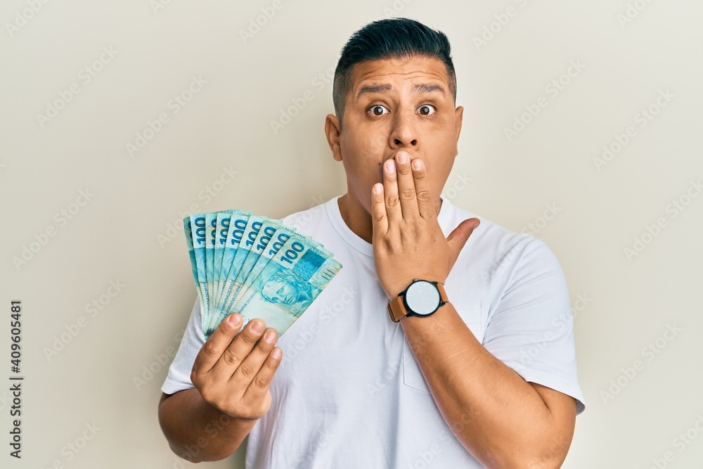 Young latin man holding 100 brazilian real banknotes covering mouth with hand, shocked and afraid for mistake. surprised expression Young latin man holding 100 brazilian real banknotes covering mouth with hand, shocked and afraid for mistake. surprised expression