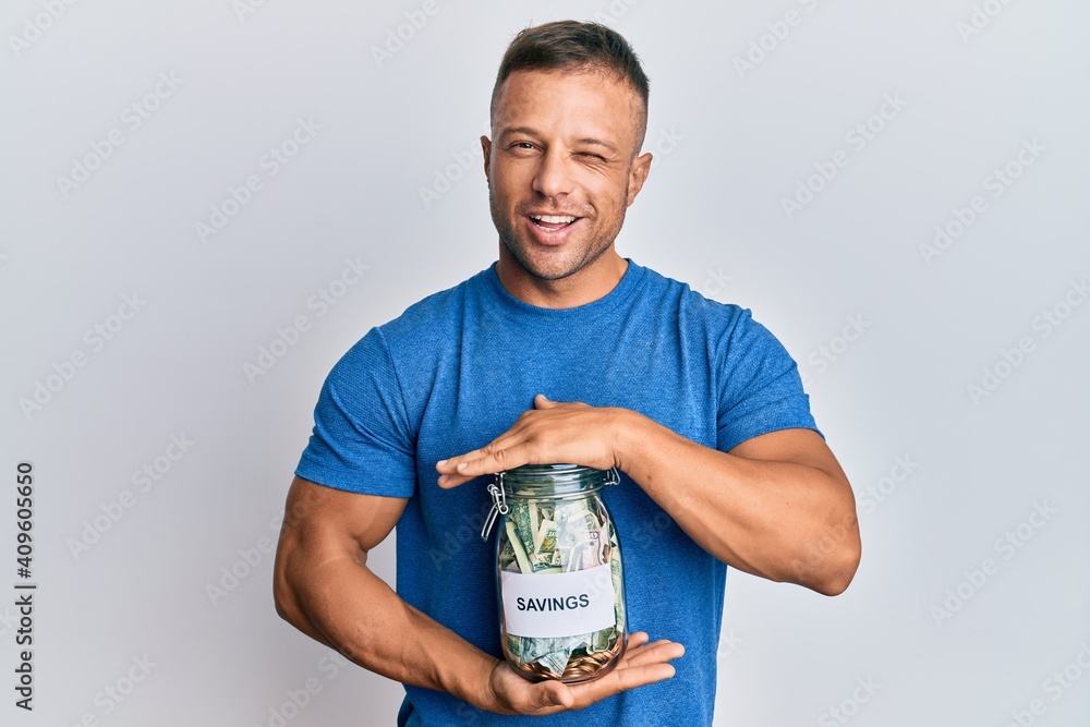 Handsome muscle man holding jar with savings winking looking at the camera with sexy expression, cheerful and happy face. Handsome muscle man holding jar with savings winking looking at the camera with sexy expression, cheerful and happy face.