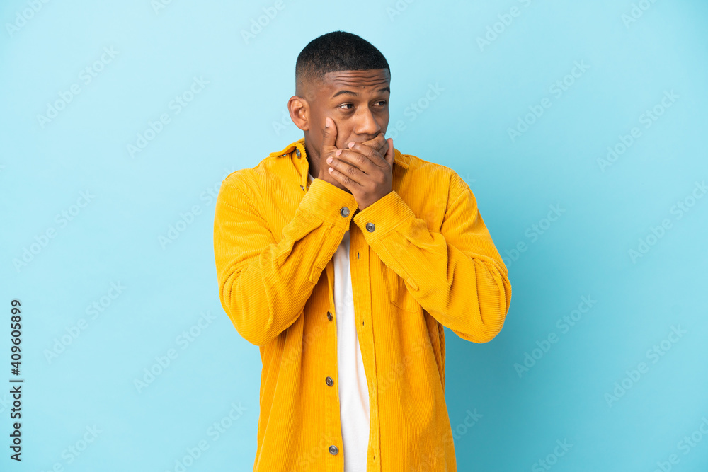 Young latin man isolated on blue background covering mouth and looking to the side