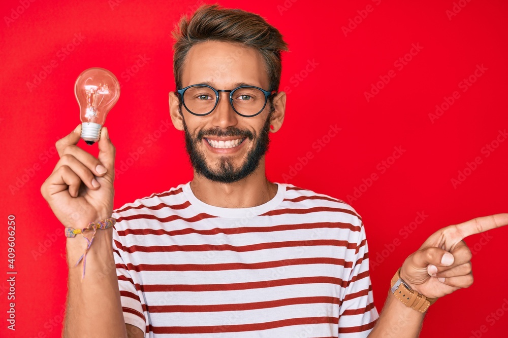 Handsome caucasian man with beard holding lightbulb for inspiration and idea smiling happy pointing with hand and finger to the side Handsome caucasian man with beard holding lightbulb for inspiration and idea smiling happy pointing with hand and finger to the side