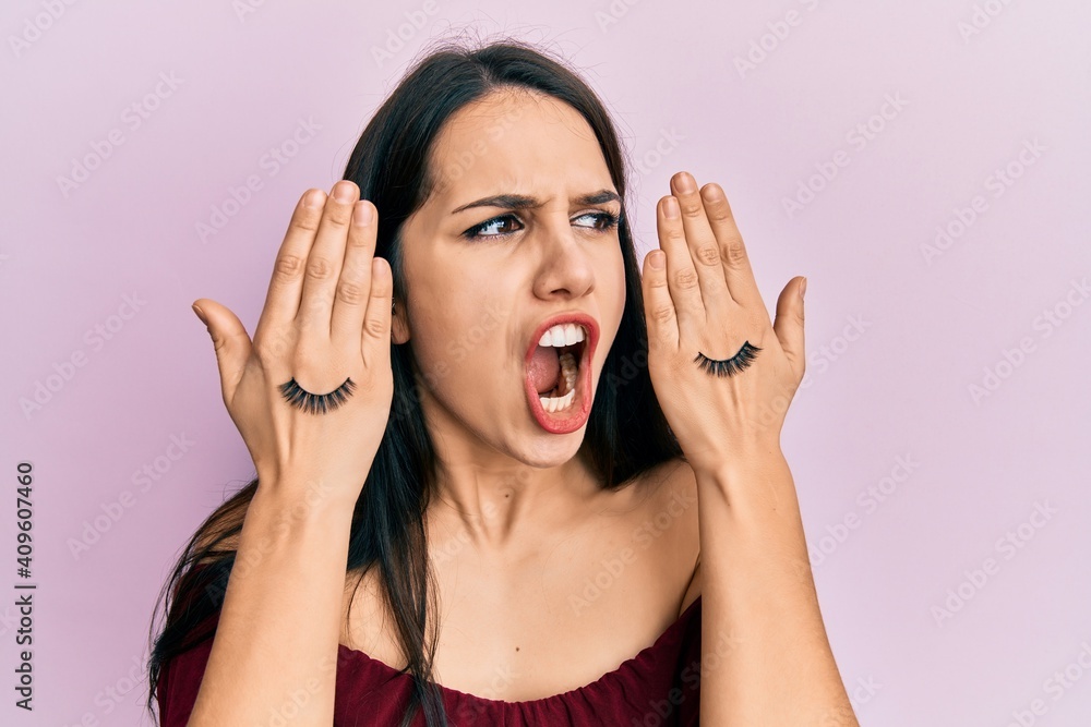 Young hispanic woman covering eyes with hands and fake lashes angry and mad screaming frustrated and furious, shouting with anger. rage and aggressive concept. Young hispanic woman covering eyes with hands and fake lashes angry and mad screaming frustrated and furious, shouting with anger. rage and aggressive concept.
