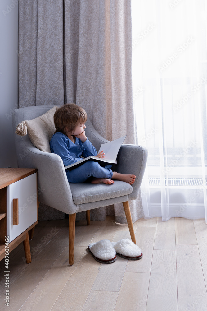 Cute preschool blonde kid reading book sitting on a cozy chair in light nordic style living room Cute preschool blonde kid reading book sitting on a cozy chair in light nordic style living room