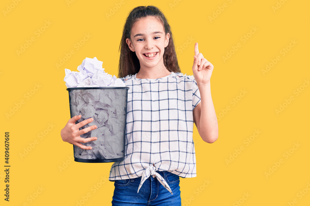 Cute hispanic child girl holding paper bin full of crumpled papers surprised with an idea or question pointing finger with happy face, number one Cute hispanic child girl holding paper bin full of crumpled papers surprised with an idea or question pointing finger with happy face, number one