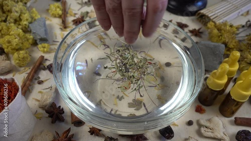 Woman's hand puts aromatic dried flowers and herbs to the glassware with a water. Preparation of natural bath for skin. Alternative medicine, organic cosmetics, aromatherapy and natural care concept.