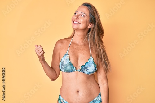 Middle age hispanic woman wearing bikini smiling with happy face looking and pointing to the side with thumb up.