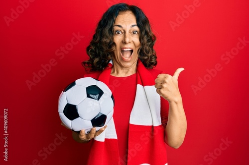 Middle age hispanic woman football hooligan holding ball pointing thumb up to the side smiling happy with open mouth