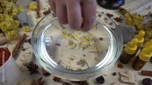 Woman's hand puts aromatic dried flowers and herbs to the glassware with a water. Preparation of natural bath for skin. Alternative medicine, organic cosmetics, aromatherapy and natural care concept.