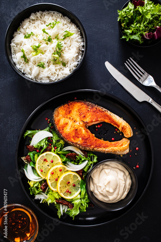 Fried salmon steak with basmati rice, mayonnaise and mix of vegetables served on black plate on wooden table 