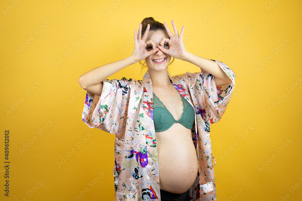 Young beautiful woman pregnant expecting baby wearing pajama over isolated yellow background doing ok gesture shocked with smiling face, eye looking through fingers Young beautiful woman pregnant expecting baby wearing pajama over isolated yellow background doing ok gesture shocked with smiling face, eye looking through fingers