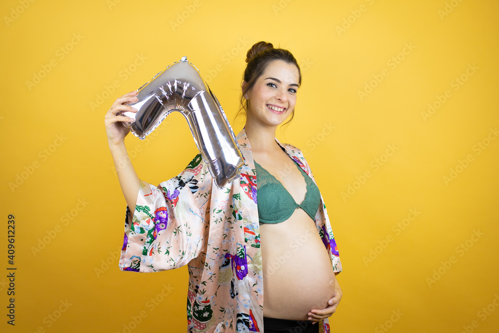 Young beautiful woman pregnant expecting baby wearing pajama over isolated yellow background holding a number seven balloon. Celebration concept Young beautiful woman pregnant expecting baby wearing pajama over isolated yellow background holding a number seven balloon. Celebration concept