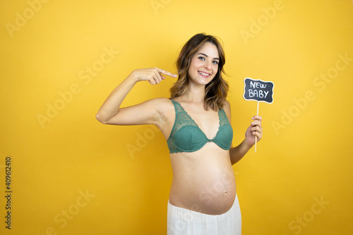 Young beautiful brunette woman pregnant expecting baby over isolated yellow background amazed and holding blackboard with new baby word message and pointing photo