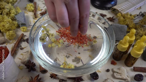 Woman's hand puts aromatic dried flowers and herbs to the glassware with a water. Preparation of natural bath for skin. Alternative medicine, organic cosmetics, aromatherapy and natural care concept.