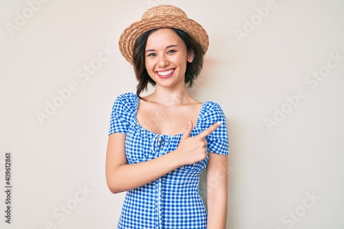 Young beautiful girl wearing summer hat smiling cheerful pointing with hand and finger up to the side