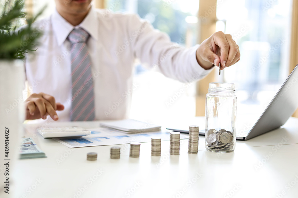 Businessman putting coins into a glass jar. He spends his monthly savings to invest in which he can grow. Ideas to make savings grow. Businessman putting coins into a glass jar. He spends his monthly savings to invest in which he can grow. Ideas to make savings grow.