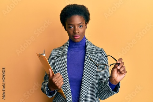 Young african american girl holding clipboard and glasses relaxed with serious expression on face. simple and natural looking at the camera.